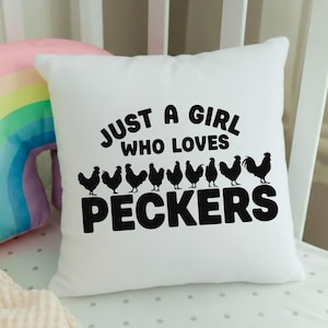 May include: White square pillow with the text "JUST A GIRL WHO LOVES PECKERS" in black. Below the text are black silhouettes of chickens. The pillow is on a white crib.