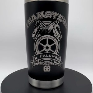 以下が含まれることがあります： シルバーの縁と蓋が付いた黒い断熱タンブラー。タンブラーには、2頭の馬と車輪のグラフィックの上に「TEAMSTERS」の文字が描かれています。グラフィックの下には「R. PALUMBO LOCAL 63」と書かれています。
