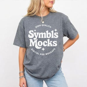 May include: A heather gray t-shirt with the text "Symbl's Mocks" in a white, retro-style font. The shirt also has the words "HIGH QUALITY" and "DIGITAL POD MOCKUPS" in white. The model is wearing blue jeans.