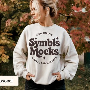 May include: Cream-colored sweatshirt with the text "Symbls Mocks" in brown, surrounded by stars and the words "High Quality" and "Digital Pod Mockups". The sweatshirt is worn by a person standing outdoors.