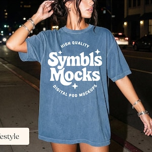 May include: A blue t-shirt with the text "Symbls Mocks" in white, with the words "High Quality" above and "Digital Pod Mockups" below. The shirt is worn by a person on a city street at night.