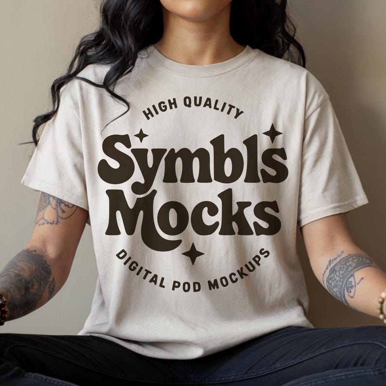 May include: A beige t-shirt featuring the words "Symbls Mocks" in a dark brown, retro font. The text is accompanied by stars and the phrases "High Quality" and "Digital Pod Mockups".