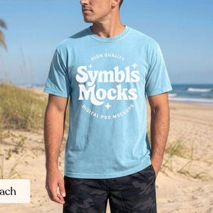 May include: A light blue t-shirt with the text "Symbl's Mocks" in white, with the words "High Quality" and "Digital Pod Mockups" above and below the main text. The shirt is worn by a person on a beach, with the ocean and sand in the background.
