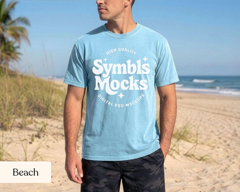 May include: A light blue t-shirt with the text "Symbl's Mocks" in white, with the words "High Quality" and "Digital Pod Mockups" above and below the main text. The shirt is worn by a person on a beach, with the ocean and sand in the background.