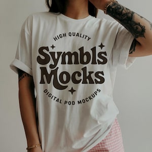 May include: A white t-shirt with rolled sleeves displaying the text "Symbls Mocks" in a brown, retro font. The shirt also features the text "High Quality" and "Digital Pod Mockups". This is a mock-up for digital products.