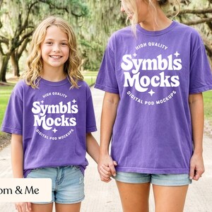 May include: Two people wearing matching purple t-shirts with the text "Symbls Mocks" in white. The shirts are paired with denim shorts. The phrase "Mom & Me" is at the bottom of the image.