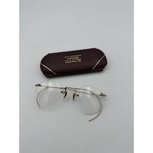 May include: Vintage rimless eyeglasses with a gold-tone metal frame and clear lenses. The glasses are next to a brown leather case with silver trim and text that reads "M.O. COCKRUM Optometrist 14 N.W. 4th Street Evansville, Ind."