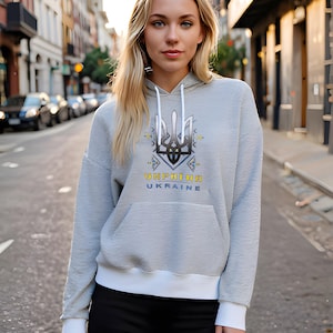 Puede incluir: Una sudadera gris con ribete blanco y un símbolo de tridente ucraniano amarillo y azul con el texto "UKRAINE" y "УКРАЇНА" impreso en la parte delantera.