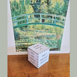 K&ouml;nnte beinhalten: Ein wei&szlig;er W&uuml;rfel mit Textaufforderungen befindet sich vor einem Gem&auml;lde einer Br&uuml;cke &uuml;ber Wasser. Der W&uuml;rfel enth&auml;lt Aufforderungen wie "Beschreibe, was du siehst" und "Welche Farben fallen dir auf?" Das Gem&auml;lde weist eine gr&uuml;ne und blaue Farbpalette auf.
