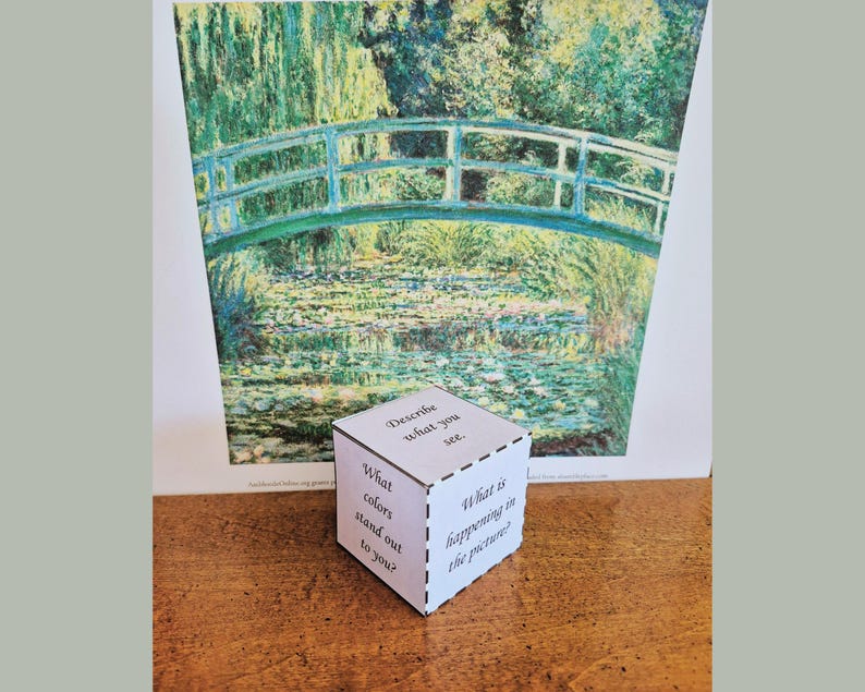 K&ouml;nnte beinhalten: Ein wei&szlig;er W&uuml;rfel mit Textaufforderungen befindet sich vor einem Gem&auml;lde einer Br&uuml;cke &uuml;ber Wasser. Der W&uuml;rfel enth&auml;lt Aufforderungen wie "Beschreibe, was du siehst" und "Welche Farben fallen dir auf?" Das Gem&auml;lde weist eine gr&uuml;ne und blaue Farbpalette auf.