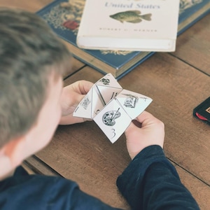 K&ouml;nnte beinhalten: Eine Person h&auml;lt einen wei&szlig;en Papier-Wahrsager mit schwarzen Illustrationen und Zahlen. Der Wahrsager zeigt Bilder einer Farbpalette, eines Geb&auml;udes und anderer Designs. B&uuml;cher sind im Hintergrund zu sehen.