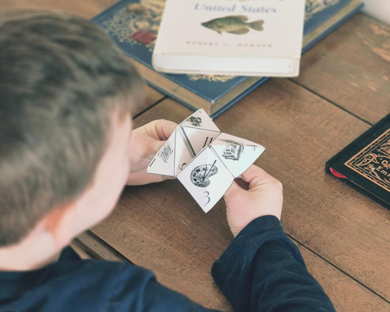 K&ouml;nnte beinhalten: Eine Person h&auml;lt einen wei&szlig;en Papier-Wahrsager mit schwarzen Illustrationen und Zahlen. Der Wahrsager zeigt Bilder einer Farbpalette, eines Geb&auml;udes und anderer Designs. B&uuml;cher sind im Hintergrund zu sehen.