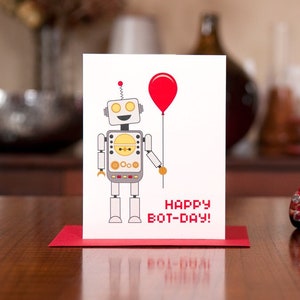 Puede incluir: Una tarjeta blanca con un robot de dibujos animados sosteniendo un globo rojo. La tarjeta dice "Happy Bot-Day!" y est&aacute; sentada en una mesa roja con un coche de juguete rojo delante.