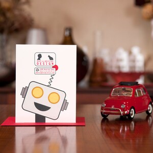 Puede incluir: Una tarjeta blanca con una ilustraci&oacute;n de un robot y el texto "Dan is George Design". La tarjeta est&aacute; sobre una mesa de madera junto a un coche de juguete rojo.