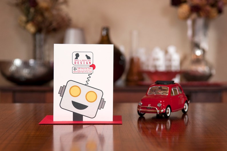 Puede incluir: Una tarjeta blanca con una ilustraci&oacute;n de un robot y el texto "Dan is George Design". La tarjeta est&aacute; sobre una mesa de madera junto a un coche de juguete rojo.