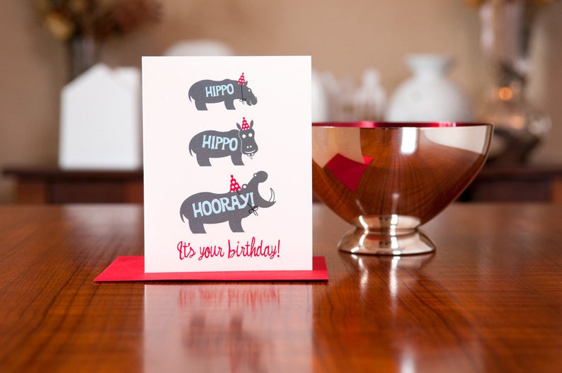 Op de afbeelding: Een witte kaart met een rode envelop. De kaart heeft drie grijze nijlpaarden met rode feestmutsen. De bovenste twee nijlpaarden zeggen "HIPPO" en het onderste nijlpaard zegt "HOORAY!" De kaart zegt "Het is je verjaardag!"