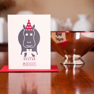 Op de afbeelding: Een rood-witte feestmuts zit bovenop een grijs silhouet van een nijlpaard. De kaart heeft de tekst "man v. george design" en bevat een websiteadres.