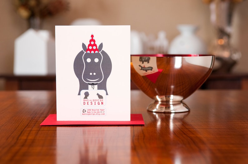 Op de afbeelding: Een rood-witte feestmuts zit bovenop een grijs silhouet van een nijlpaard. De kaart heeft de tekst "man v. george design" en bevat een websiteadres.