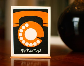 Puede incluir: Un teléfono retro de estilo negro y naranja con un dial rotatorio y el texto "Give Me a Ring!" impreso en la parte delantera.
