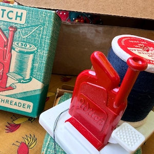 May include: A red plastic Witch needle threader on a white base, positioned next to a spool of dark blue thread. The original green and white cardboard box is visible, featuring an illustration of the threader and the text Witch Needle Threader.