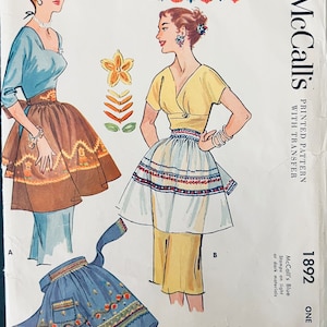 Op de afbeelding: Een vintage naaipatroon voor twee korte schorten. Het patroon is voor McCall's 1892 en is gedrukt op blauw papier met een transfer. De schorten worden getoond op twee vrouwen die ze dragen. Een schort is bruin met een witte rand en de andere is wit met een rode, blauwe en witte rand. Het patroon bevat instructies voor het maken van de schorten en een transfer voor de borduurwerk.