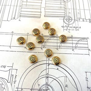 May include: A collection of 12 gold-colored metal beads with a spiral design. The beads are scattered on a white surface with a blueprint pattern.