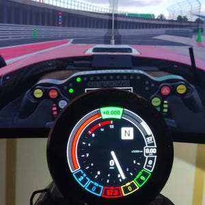 Peut inclure: Gros plan sur une jauge numérique noire affichant des données de course colorées. La jauge est placée devant le tableau de bord d'une voiture de course et un circuit simulé.