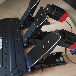 Op de afbeelding: Zwarte racepedalen met zilveren accenten en het "MOZA RACING" logo. De pedalen zijn gemonteerd op een houten basis. De pedaalset bevat drie pedalen en twee zwart-rode modules.