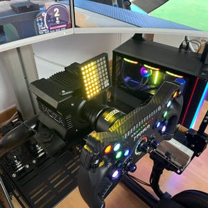Könnte beinhalten: Ein schwarzes Rennsimulator-Lenkrad mit Carbonfaser-Optik, bunten Tasten und dem Moza-Logo. Es ist an einer Direct-Drive-Basis mit einem hellgelben LED-Anzeigefeld montiert und befindet sich vor einem Computergehäuse mit RGB-Beleuchtung.