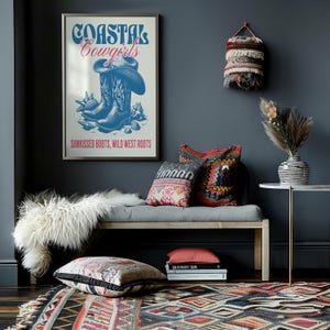 May include: A framed poster with the text "Coastal Cowgirls" in blue and red lettering. The poster features a pair of blue cowboy boots with seashells and the text "Sunkissed Boots, Wild West Roots". The poster is hanging on a dark gray wall above a gray bench with pillows and a white faux fur throw.