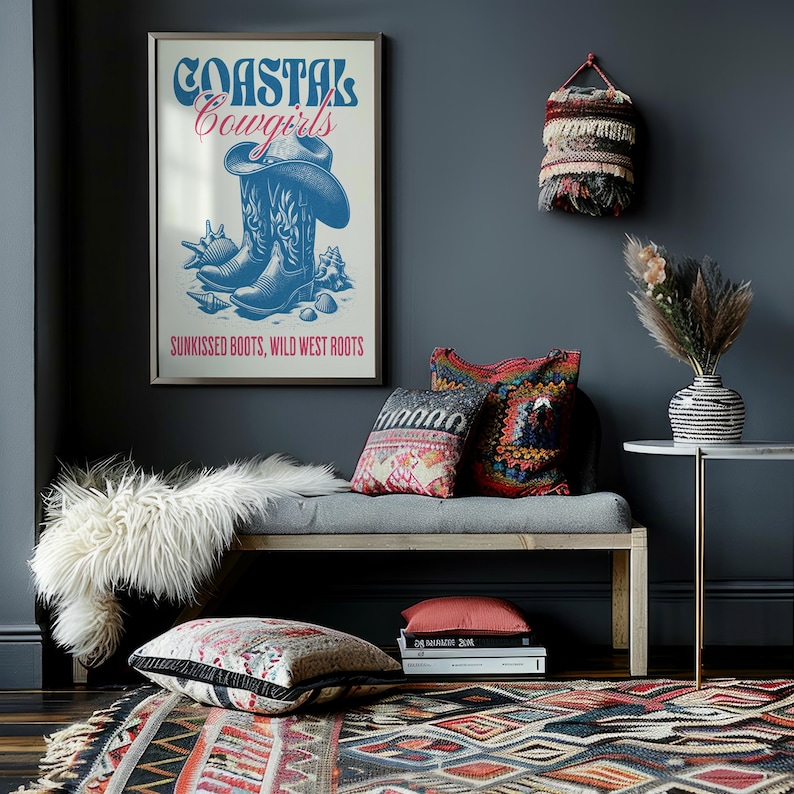 May include: A framed poster with the text "Coastal Cowgirls" in blue and red lettering. The poster features a pair of blue cowboy boots with seashells and the text "Sunkissed Boots, Wild West Roots". The poster is hanging on a dark gray wall above a gray bench with pillows and a white faux fur throw.