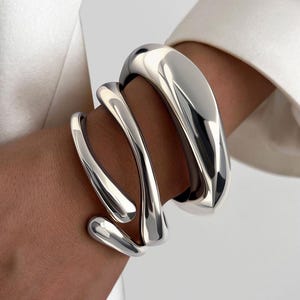 May include: A polished silver bracelet with a fluid, organic design. The bracelet wraps around the wrist with three interconnected, curved bands. The smooth, reflective surface catches the light, creating highlights and shadows. A modern piece of jewelry.