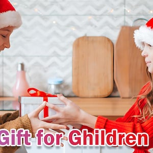 May include: Two children wearing Santa hats exchange a gift wrapped in red and white paper with a gold bow. The gift is on a table with other wrapped gifts in the background. The text "The Great Gift for Children" is at the bottom of the image.