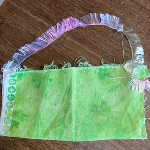 May include: A green fabric square with a white border and numbers printed on it. The fabric is fringed with a pink and white trim.