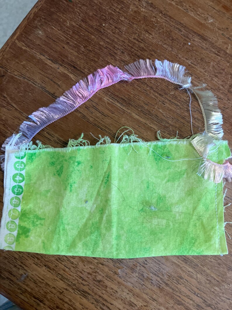 May include: A green fabric square with a white border and numbers printed on it. The fabric is fringed with a pink and white trim.