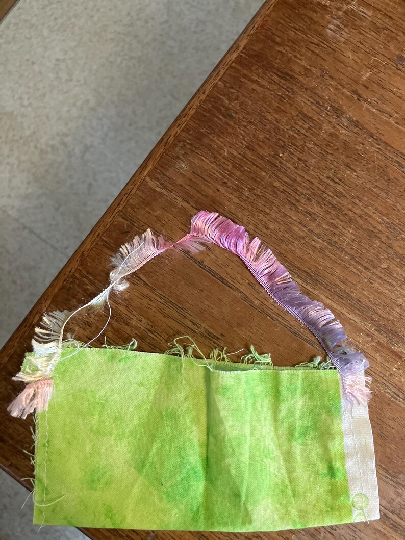 May include: A green fabric scrap with a frayed edge and a pink, purple and white fringe trim.