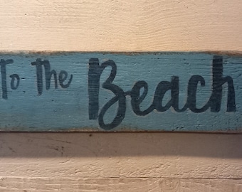 May include: A rustic, arrow-shaped wooden sign painted light blue with the words "To the Beach" in black script. The sign has a distressed finish, suggesting a vintage or coastal theme. It is mounted on a light-colored wooden background.