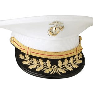 Może przedstawiać: Biały kapelusz galowy z złotą i czerwoną wstążką oraz złotym orłem. Kapelusz ma czarny rondel z wyszywaną złotą wieńcem laurowym.