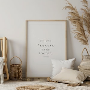 May include: A framed print with a white background and black text that reads "We love because he first loved us. 1 John 4:19". The print is hanging on a white wall with a wicker basket and a throw blanket in the foreground.