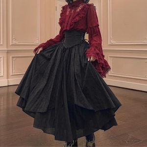 Pode incluir: Uma blusa de manga comprida em vermelho escuro com acabamento em renda, detalhes de babados e gola alta, combinada com um corpete estilo espartilho preto e uma saia preta volumosa até o chão com painéis em camadas e silhueta fluida.