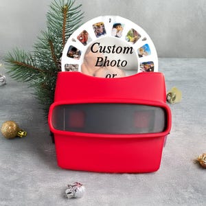 Könnte beinhalten: Ein rotes View-Master-Spielzeug mit einer weißen Foto-Rolle. Die Rolle zeigt mehrere kleine Fotos. Der Text "Custom Photo or" ist auf der Rolle aufgedruckt. Das Spielzeug steht auf einer grauen Oberfläche mit Weihnachtsdekorationen.