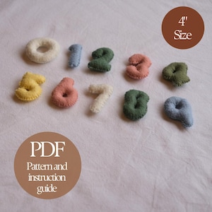 May include: A selection of felt numbers in assorted colors, such as cream, blue, green, pink, and yellow. Each number is approximately 10 cm in size. The image also includes text that says "PDF Pattern and instruction guide."