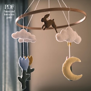 May include: A handmade baby mobile featuring a wooden ring with felt shapes. The mobile includes a brown bunny, white clouds, blue and yellow flowers, and a yellow crescent moon. The text "PDF Pattern and instruction guide" is visible.