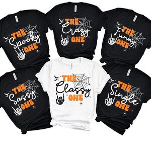 May include: A collection of black and white t-shirts with Halloween-themed designs. Each shirt features a spiderweb, bats, and the phrase "The Spooky One," "The Crazy One," "The Funny One," "The Sassy One," "The Classy One," or "The Single One."