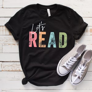 May include: Black t-shirt featuring the text "Let's READ" in a rainbow of colours. The shirt is styled with grey canvas trainers with white laces. The shirt is knotted at the bottom.