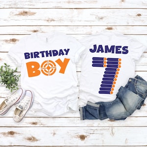 May include: Two white t-shirts with a birthday theme. One shirt says "Birthday Boy" with an orange target graphic. The other shirt says "JAMES" with a blue number 7 graphic made of orange circles.