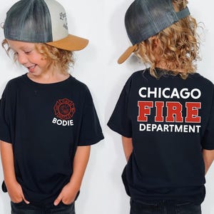 May include: Black t-shirt featuring a Chicago Fire Department design. The front of the shirt displays a red and white fire department emblem with the name "Bodie". The back of the shirt has the words "Chicago Fire Department" in white and red.
