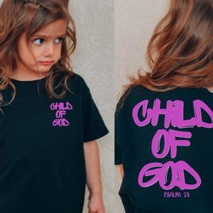 May include: Black t-shirt with the words "CHILD OF GOD" in a purple, graffiti-style font on the front and back. The back also includes "PSALM 23". The shirt is worn by a young child.
