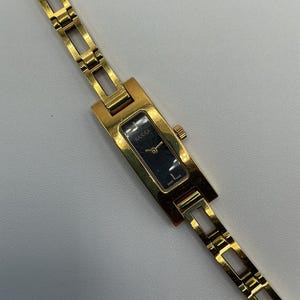 Puede incluir: Un reloj de pulsera rectangular en tono dorado con una esfera oscura con el logotipo de Gucci y una pulsera de eslabones. La esfera muestra el texto Gucci y Swiss Made. La caja y la correa de metal tienen un acabado pulido.