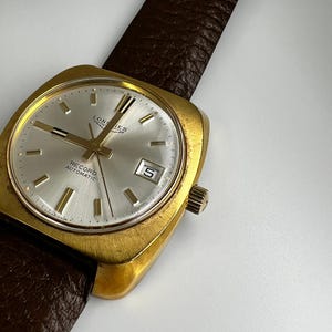 Op de afbeelding: Een vintage goudkleurig Longines Record Automatic horloge met een zilveren wijzerplaat en een bruine leren band. Het horloge heeft een datumvenster op 3 uur. Het horloge heeft een vierkante kast en gouden wijzers.